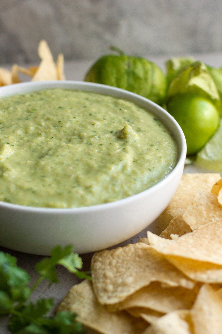 creamy salsa verde in a bowl surrounded by avocados, chips and tomatillos