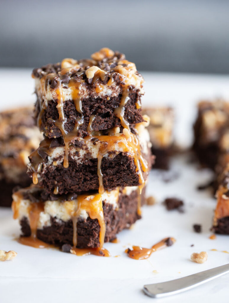 three turtle cheesecake brownies stacked high with caramel sauce dripping down