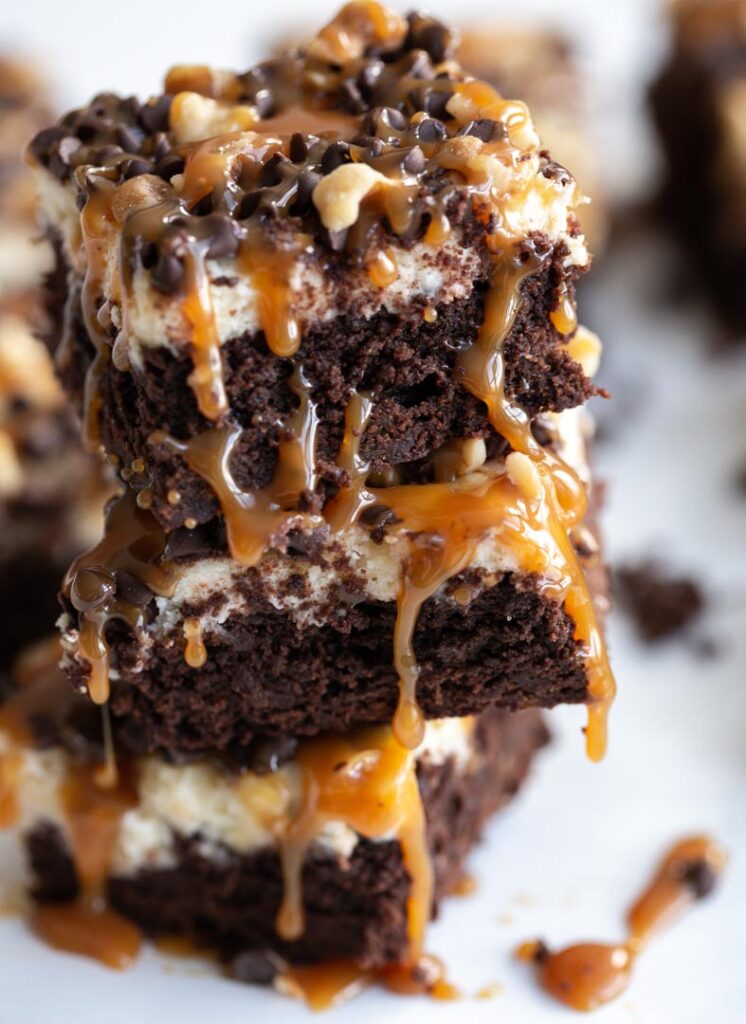 three turtle cheesecake brownies stacked on top of each other with caramel sauce dripping down