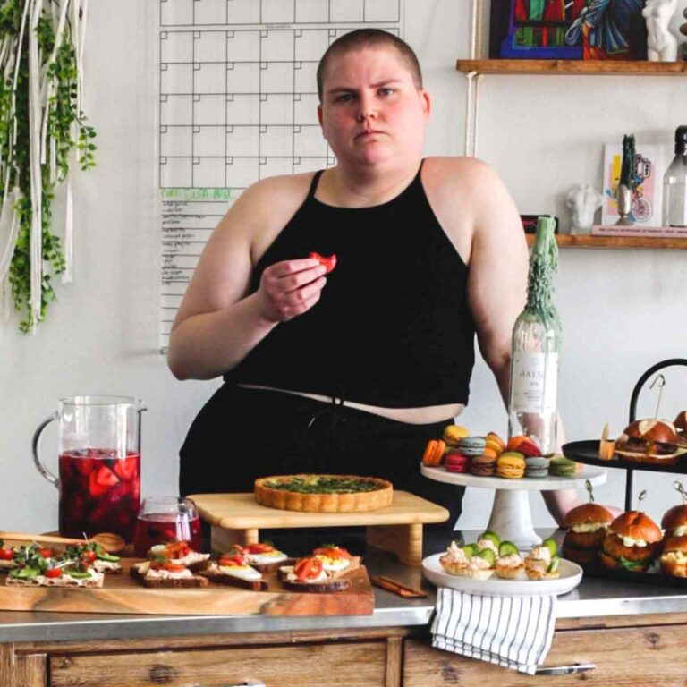 Person in a black shirt eating a macaron and looking at the camera while there is sangria, toast, quiche, sliders, and phyllo cups on the table in front of them