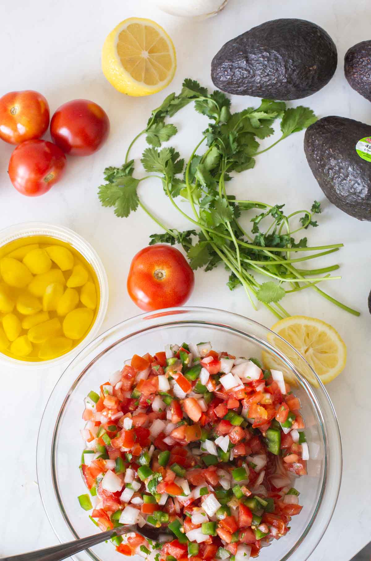 top down view of pico de gallo in a glass bowl with a spoon and tomatoes, lemons, cilantro and avocados in the background