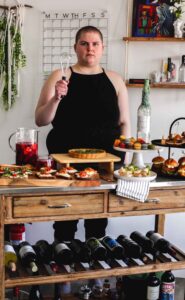 A person holding a whisk standing behind a big table of food including: quiche, toast, macarons, sliders, and sangria